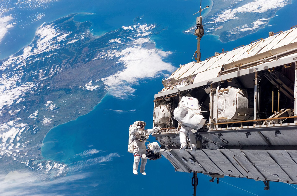 Image of the ISS above Earth with astronauts in space suits surrounding it.