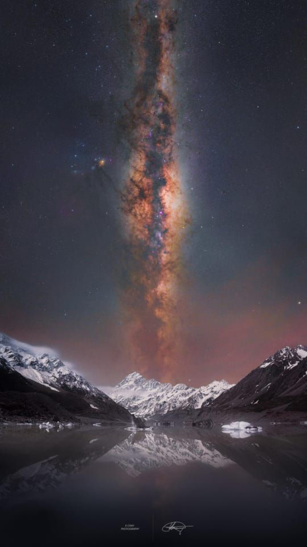 A detailed view of the Milky Way rises above an icy lake and snow capped mountains.